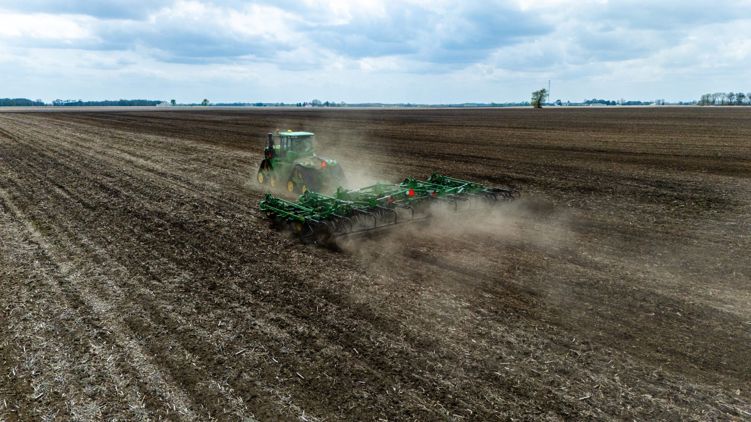 Wide drone shot of tractor and cultivator working vast Illinois farmland with flat horizon and overcast sky