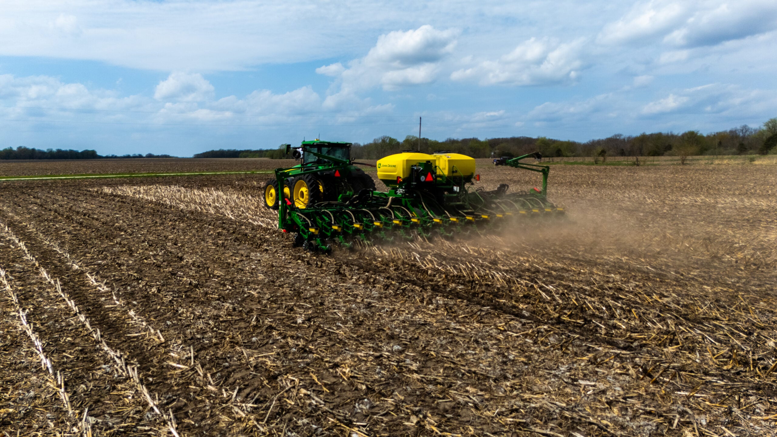 Drone photo from behind John Deere planter showing row units planting into corn stubble with dust and dramatic sky in West Central Illinois