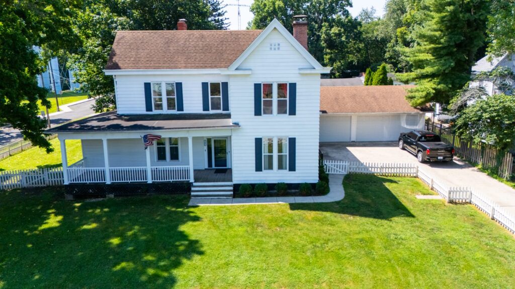 Front elevation drone photo of the white farmhouse at 1405 W College Ave, Jacksonville IL with covered porch, black shutters, and American flag