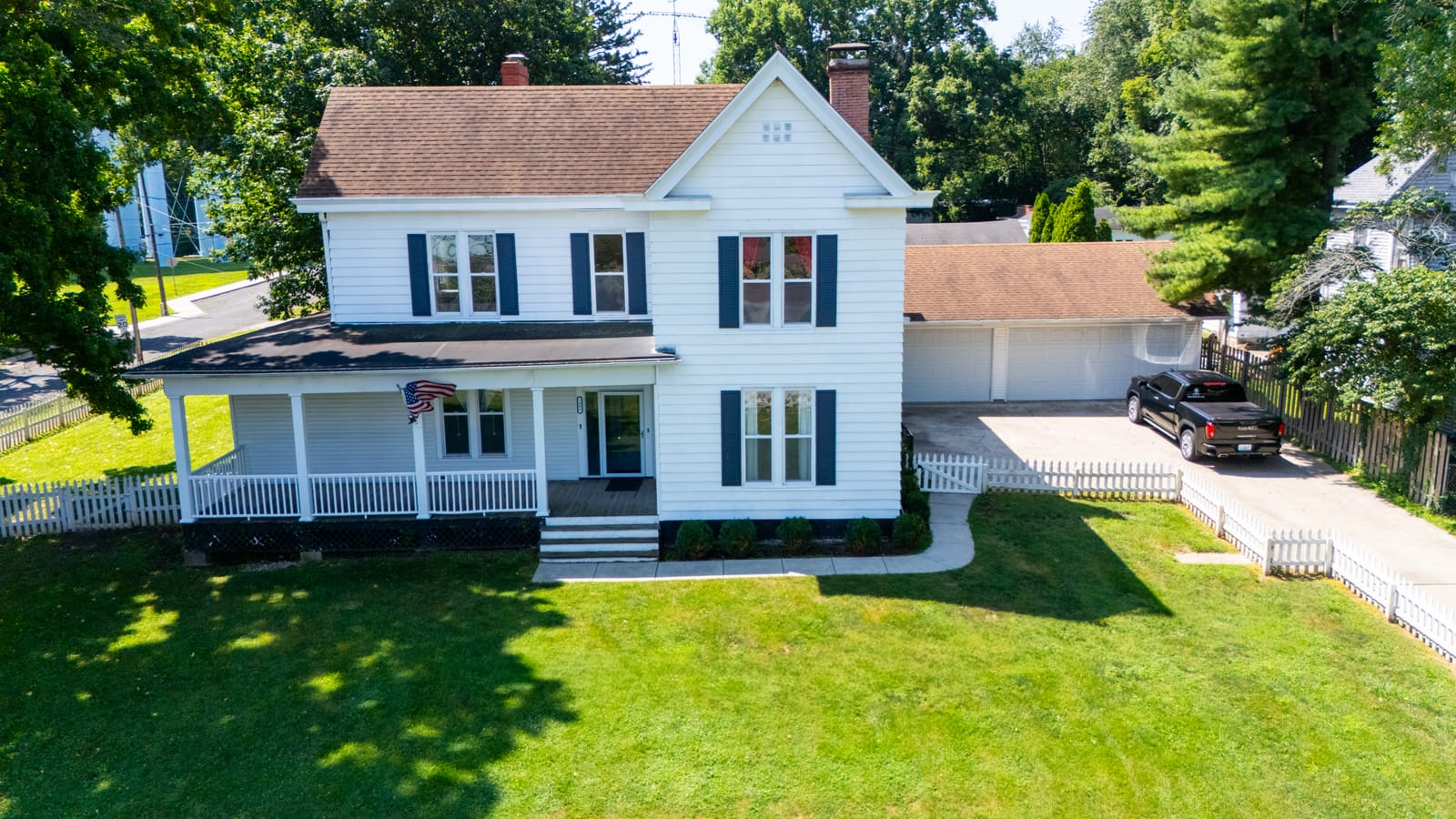 Front elevation drone photo of the white farmhouse at 1405 W College Ave, Jacksonville IL with covered porch, black shutters, and American flag