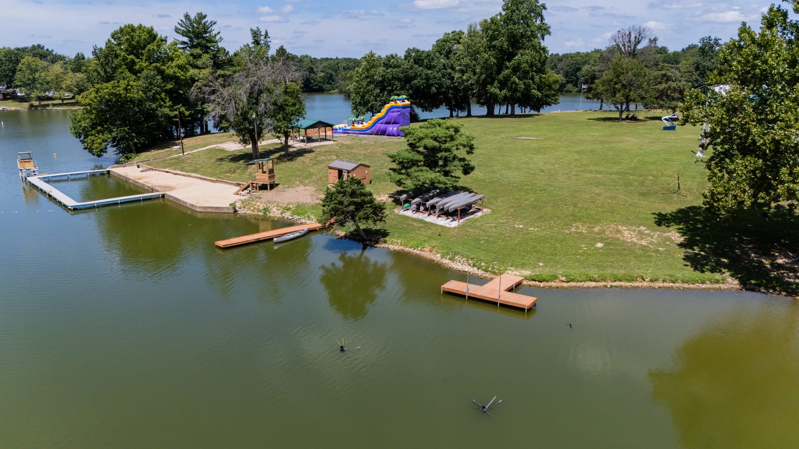 Aerial drone photo of the Western Illinois Youth Camp waterfront peninsula on Lake Jacksonville with swim docks, waterslide, and shaded picnic areas