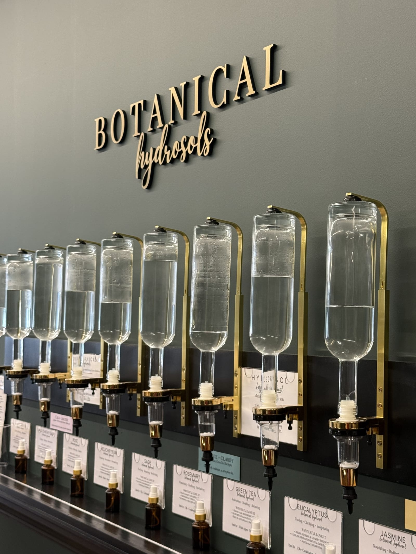 Row of glass gravity dispensers on the Custom HYDR8 Blend wall at Cheryl Kelly & Co. in Jacksonville Illinois, where guests choose up to three botanical hydrosols for a custom facial mist