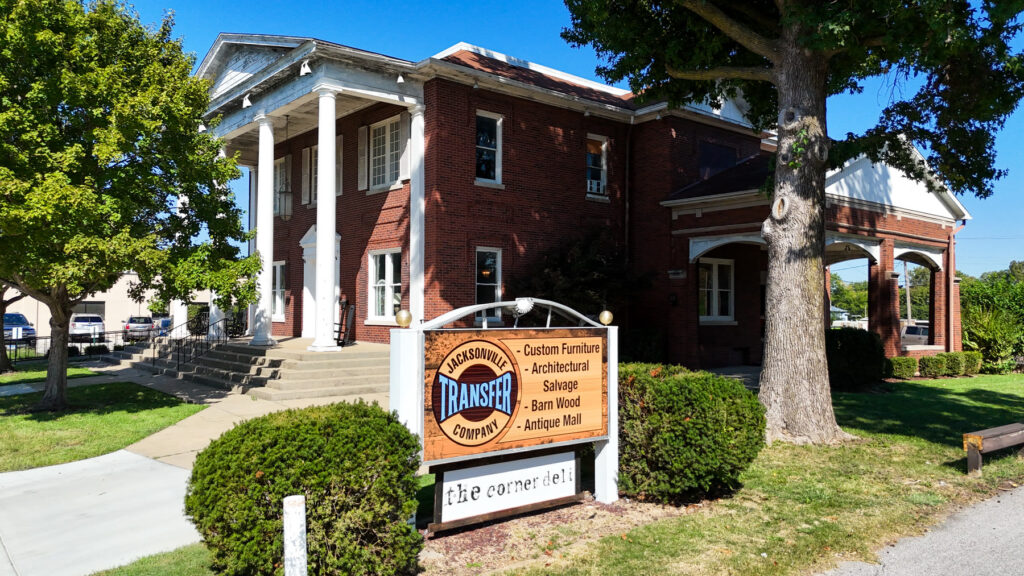 Low aerial drone photo of the Jacksonville Transfer Company sign listing custom furniture, architectural salvage, barn wood, and antique mall, in front of the red brick Gillham House with white columns at 326 W State St in Jacksonville, Illinois